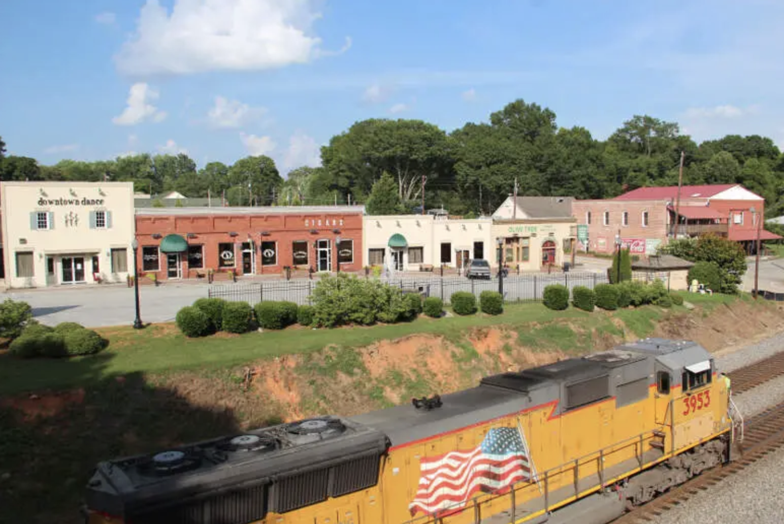 The image shows a small town with shops and a train passing by. It features trees, a blue sky, and a landscaped area.