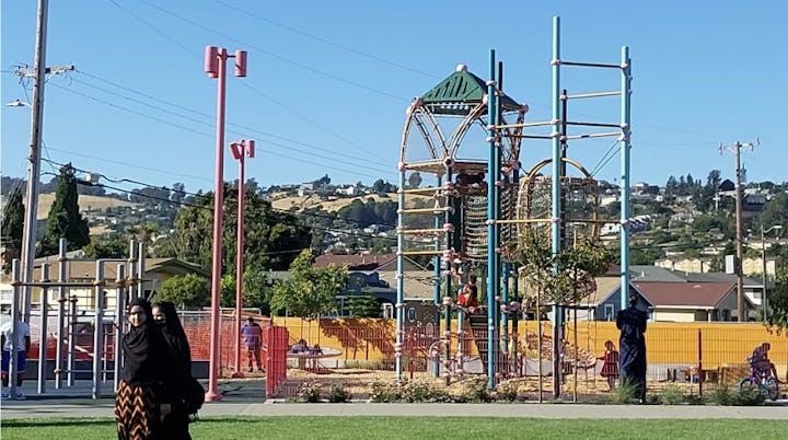 A playground with a climbing structure, people, and a scenic hill backdrop.