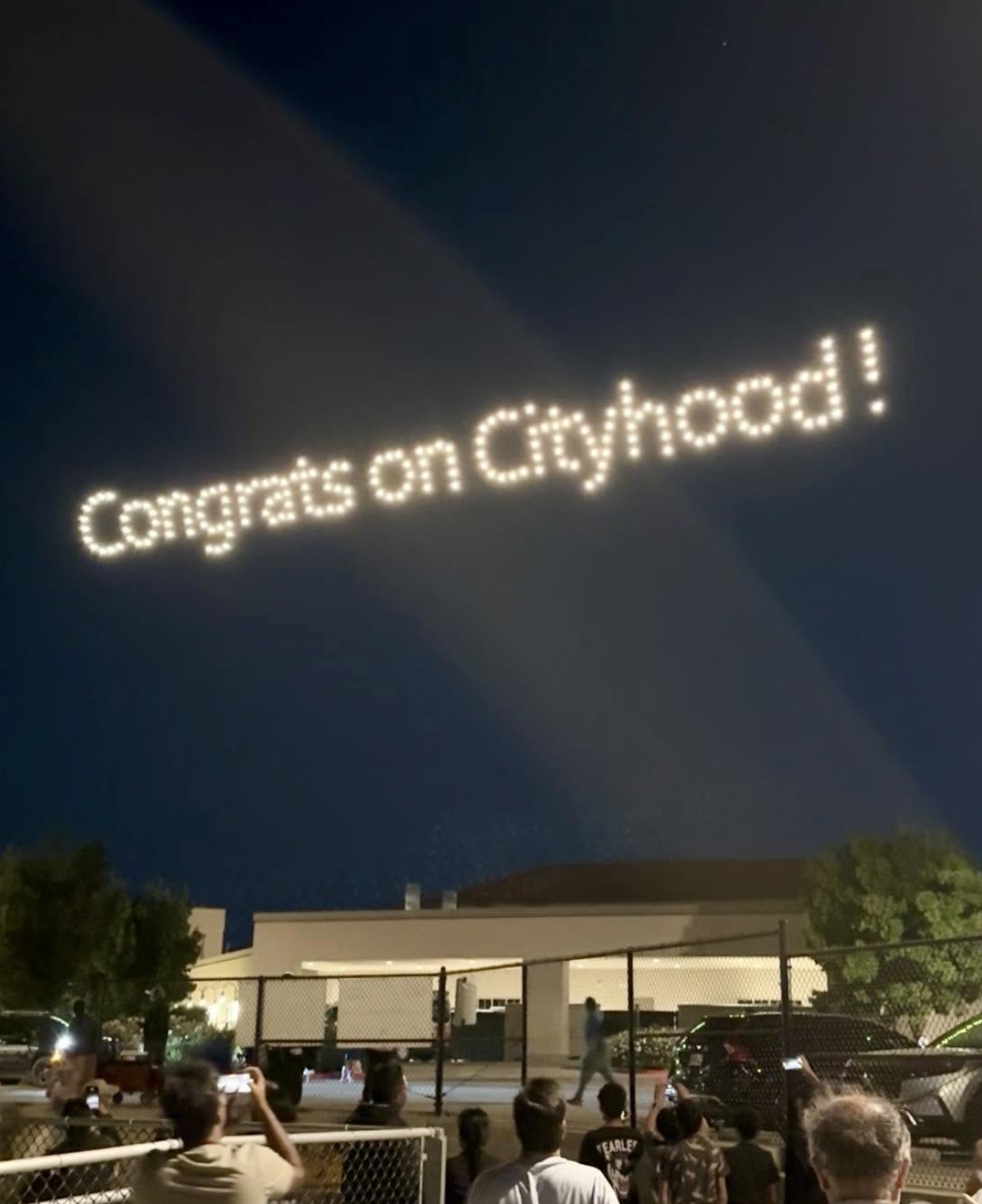 People watching a drone light display in the sky reading "Congrats on Cityhood!"