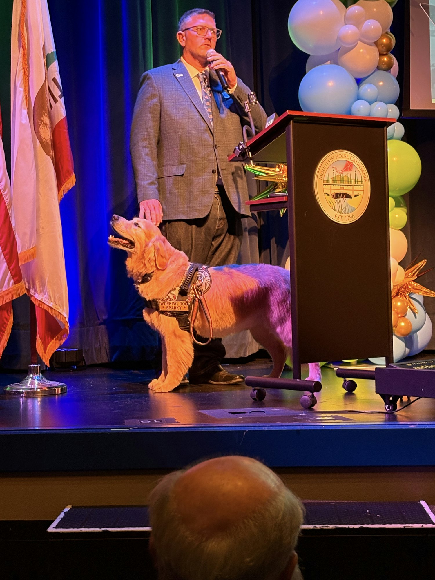 A man in a suit speaks at a podium with balloons and flags. A service dog is beside him.