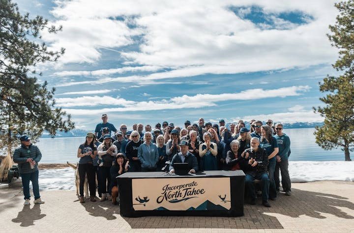 A large group of people gathered outdoors by a lake, celebrating the proposed incorporation of North Tahoe, with a table display.