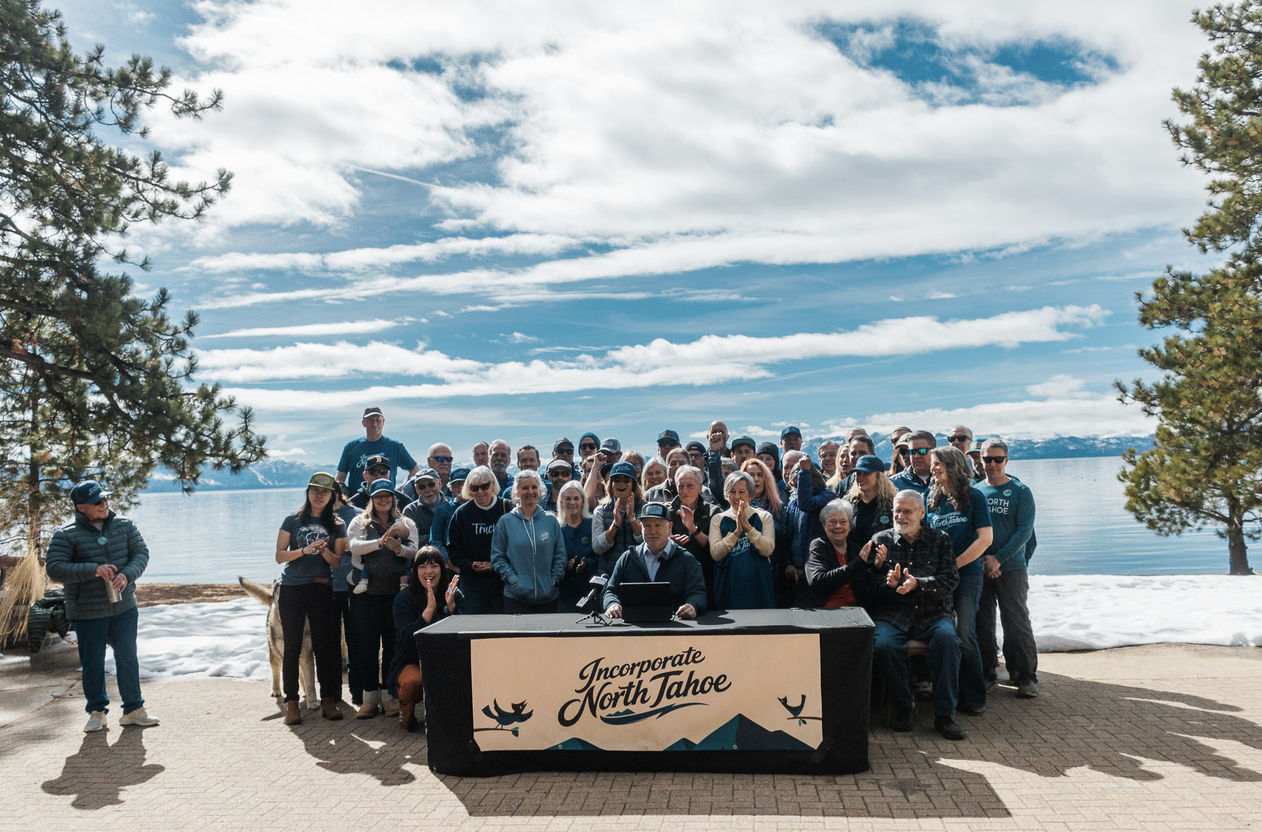 A large group of people gathered outdoors by a lake, celebrating the proposed incorporation of North Tahoe, with a table display.