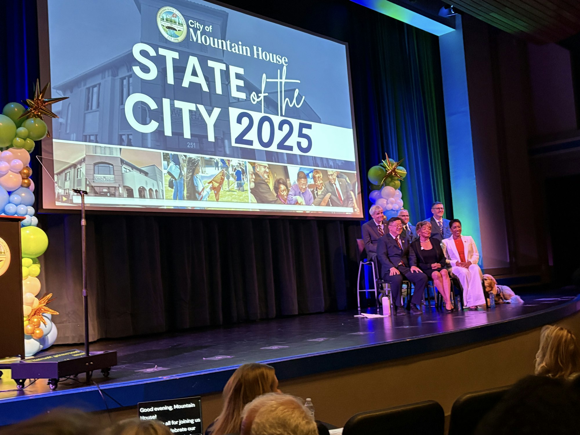 People on stage at the "State of the City 2025" event with a large screen and balloons in the background.