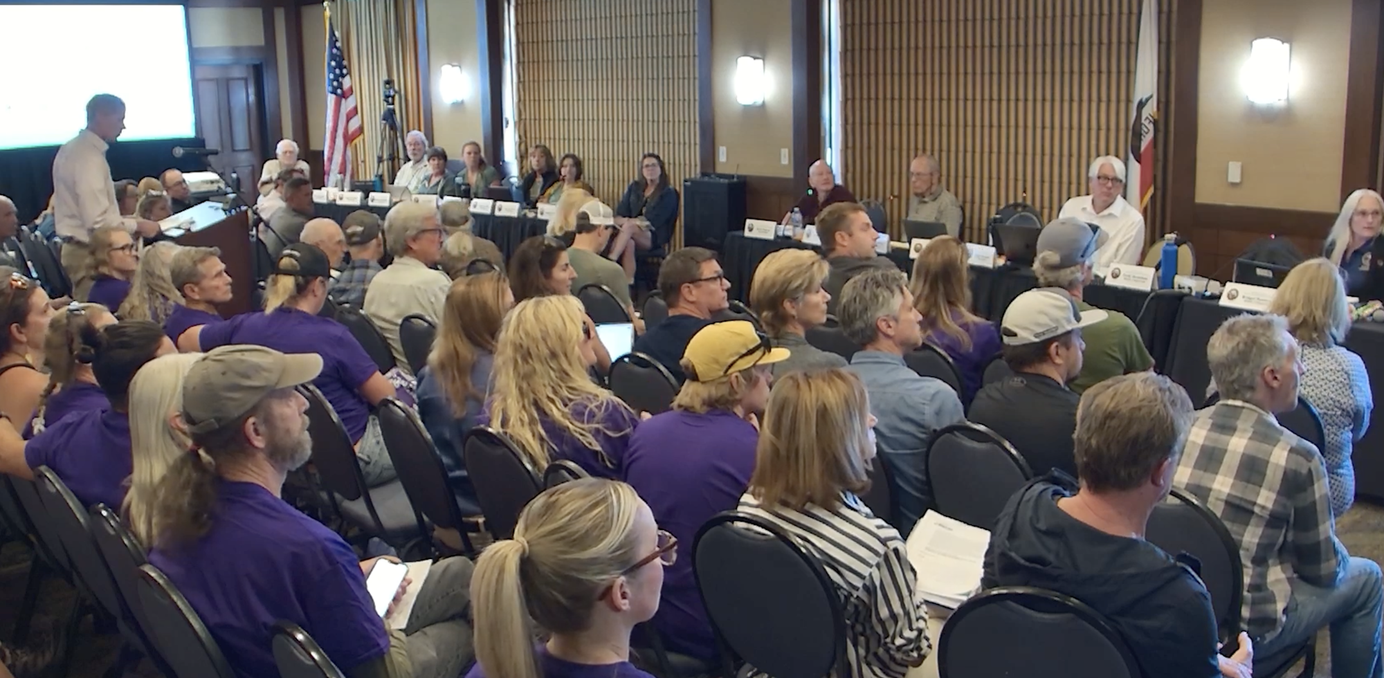 A crowded public meeting with attendees in purple shirts and panelists at the front.