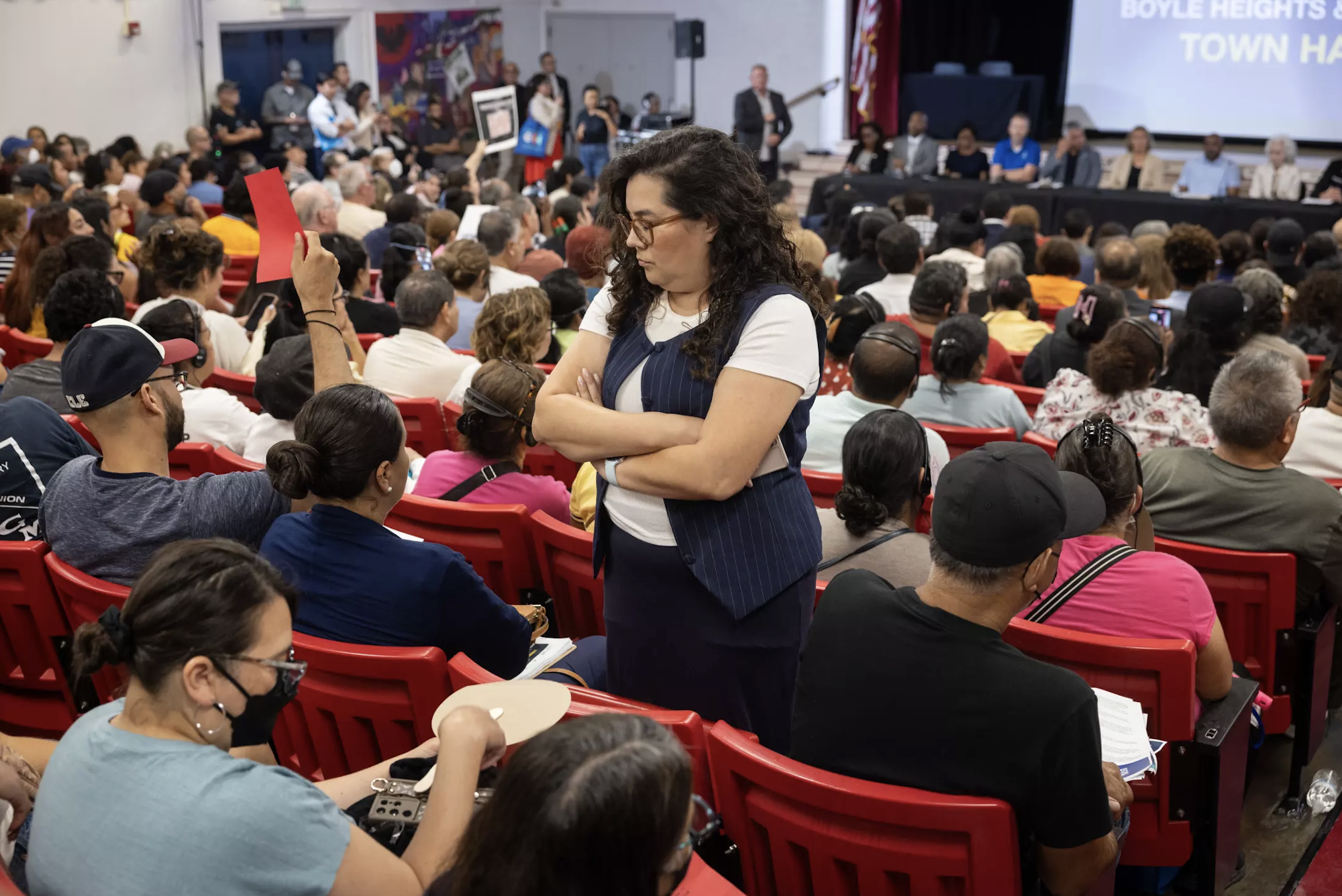 A crowded town hall meeting in Boyle Heights, with attendees engaged and a speaker looking serious in the foreground.