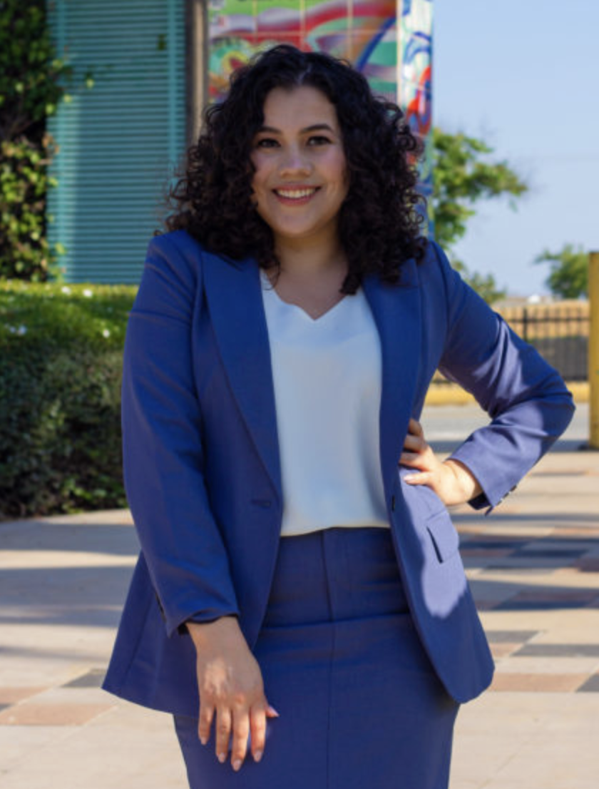 A smiling woman in a blue suit standing outdoors with her hand on her hip.