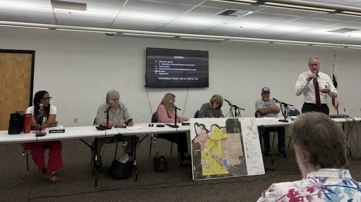 A panel discussion with six people, one speaking, agenda on screen, and a map displayed on an easel.