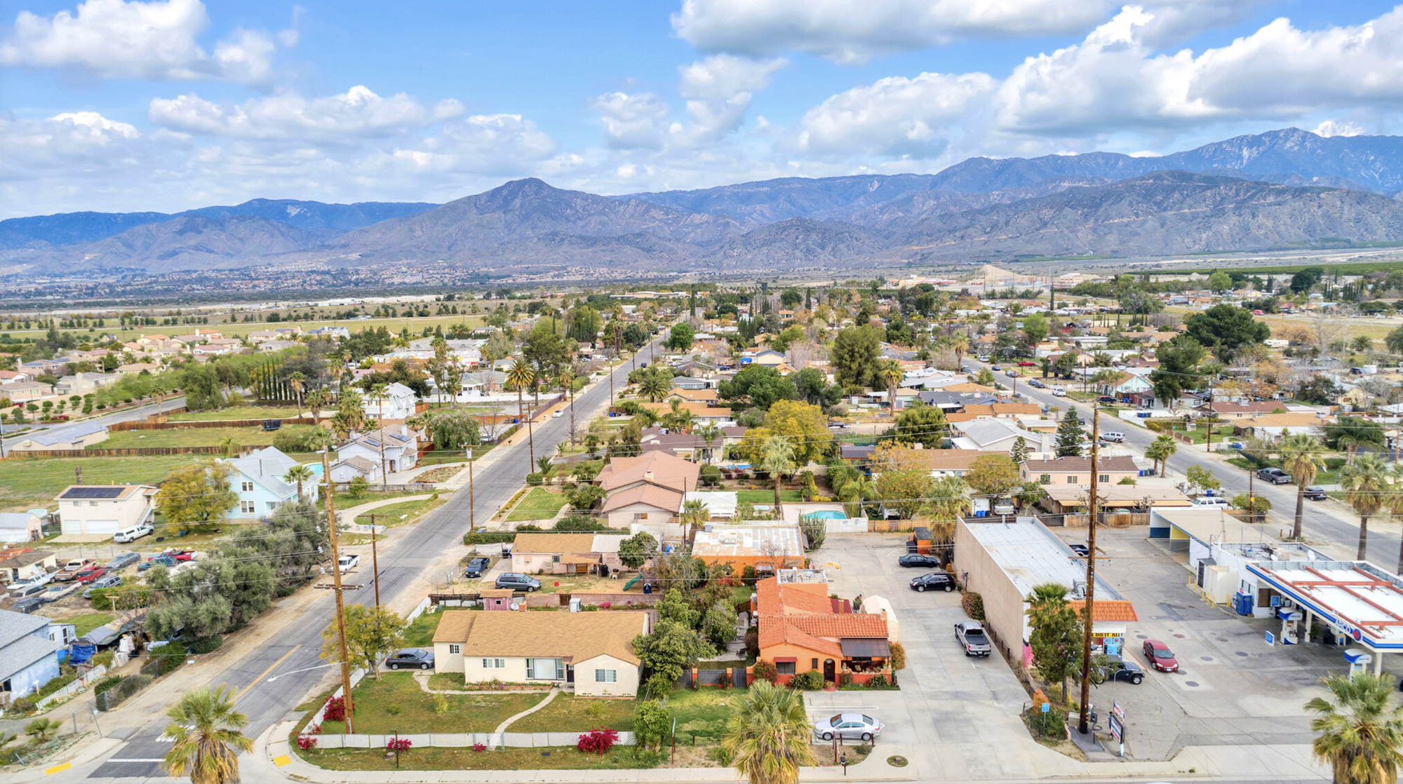 Suburban landscape with houses, trees, and a gas station in the foreground, mountains in the background under a blue sky with clouds.