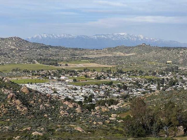 Aerial view of a hilly landscape with a small town, green fields, and distant snow-capped mountains under a cloudy sky.