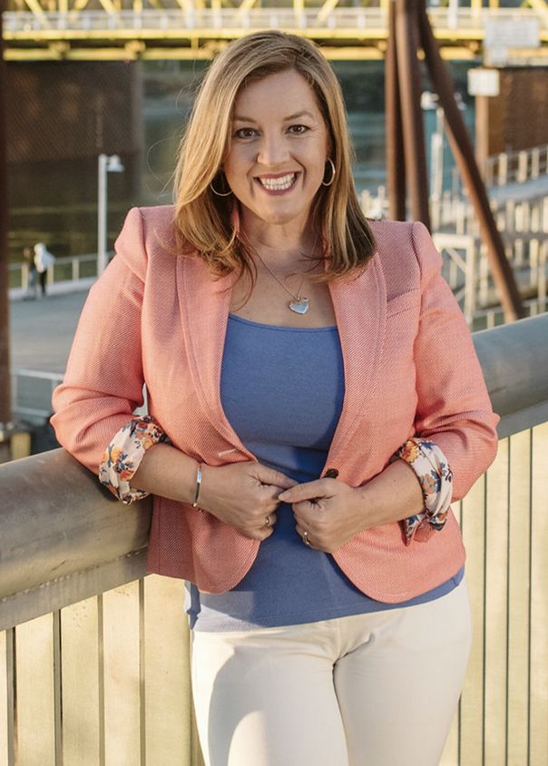 A smiling Senator Ashby in a pink blazer and blue top, standing by a railing outdoors.