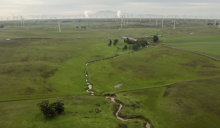 Aerial view of a green landscape with a winding stream, rolling hills, and wind turbines in the background.