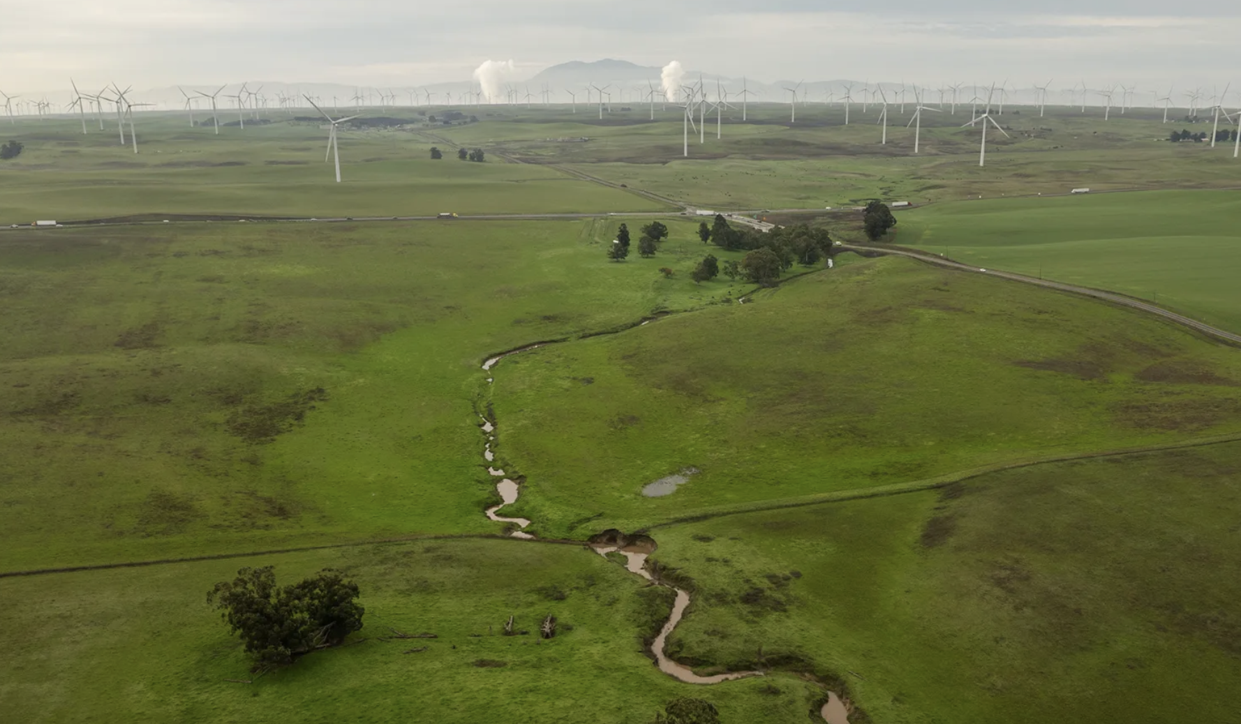 Aerial view of a green landscape with a winding stream, rolling hills, and wind turbines in the background.