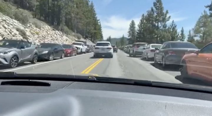 A long line of parked cars on a narrow road surrounded by trees and rocky terrain, likely in a scenic area.