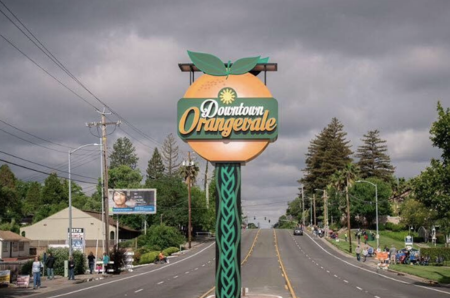 A sign for "Downtown Orangevale" designed like an orange, on a street with trees and a cloudy sky.