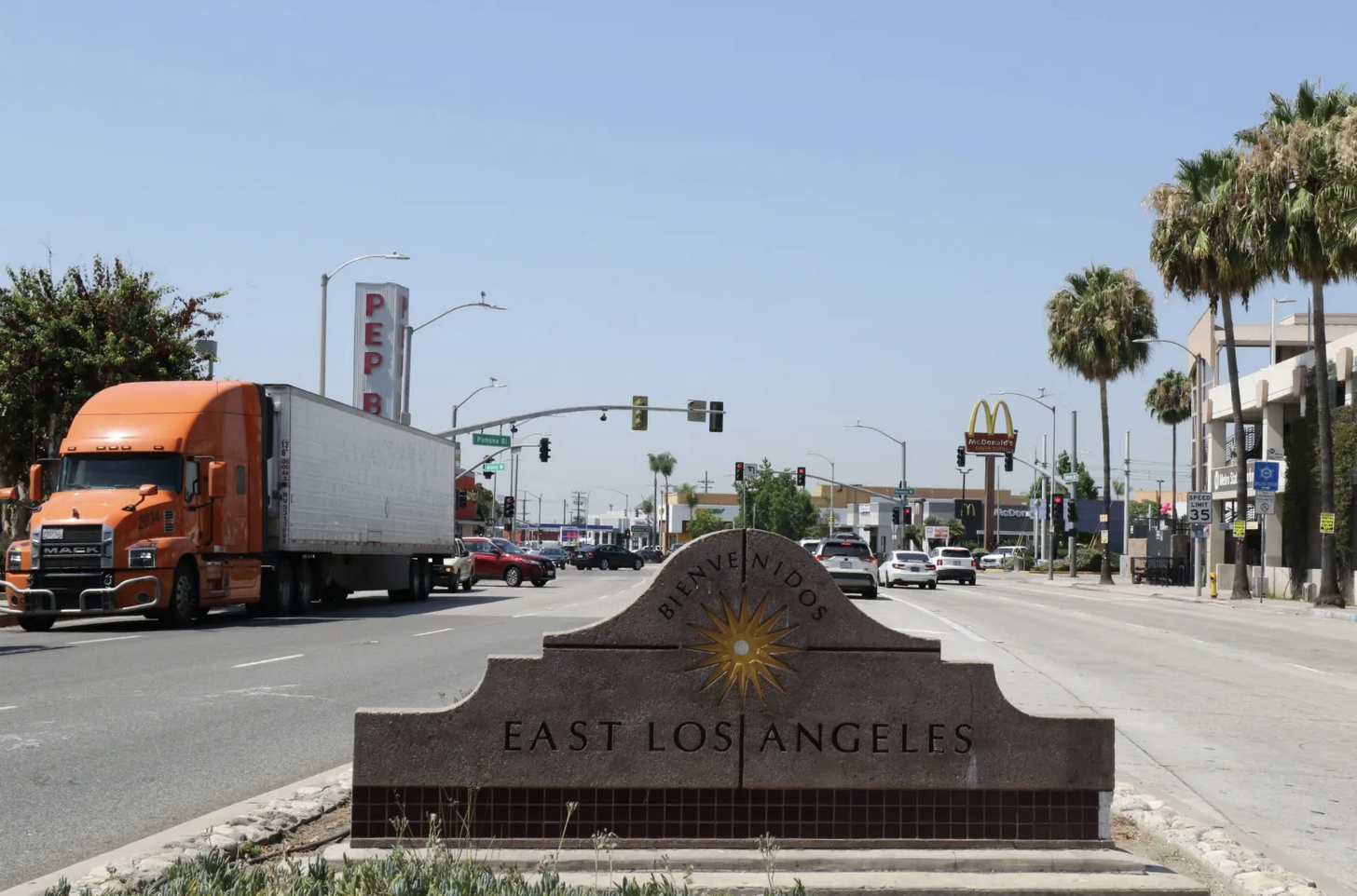 A "Bienvenidos East Los Angeles" sign by a busy street with cars, trucks, palm trees, and a McDonald's in the background.