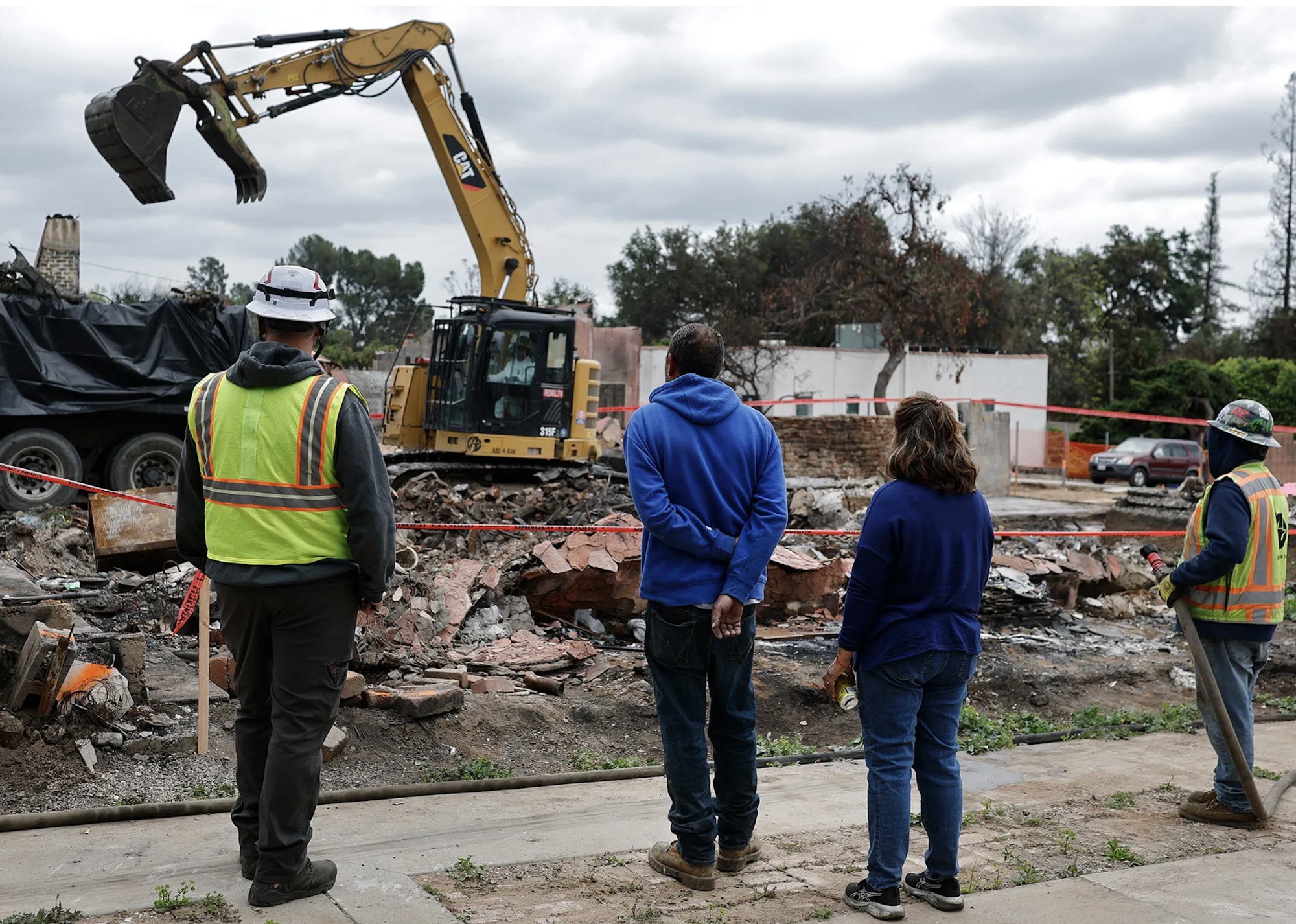 A group of people observes a demolition site with heavy machinery clearing rubble. The atmosphere looks somber and reflective.