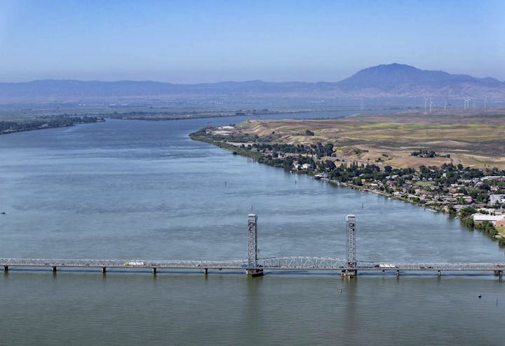 A scenic view of a river and bridge, with hills and a distant mountain backdrop under a clear blue sky.