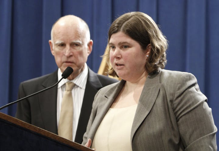 Two people at a podium, a woman speaking and a man looking on, both in professional attire.