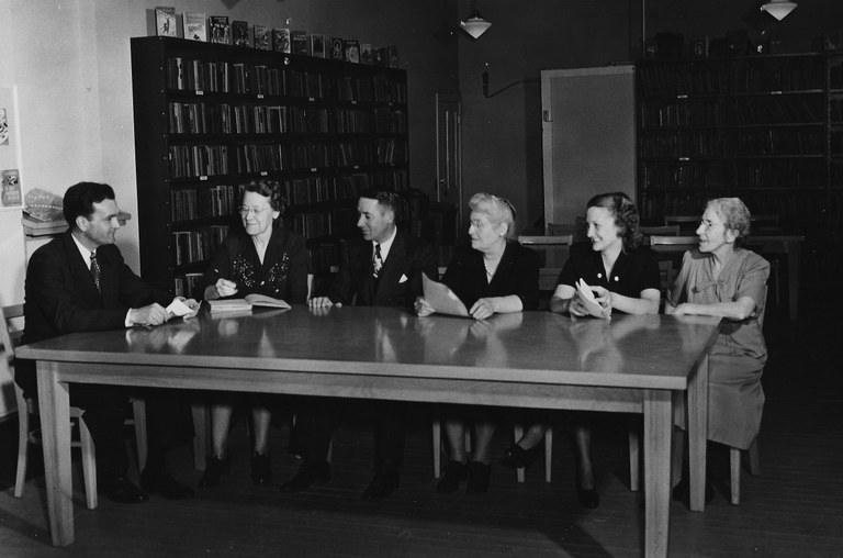 May contain: book, publication, desk, furniture, table, adult, male, man, person, chair, accessories, formal wear, tie, indoors, library, clothing, footwear, shoe, and glasses