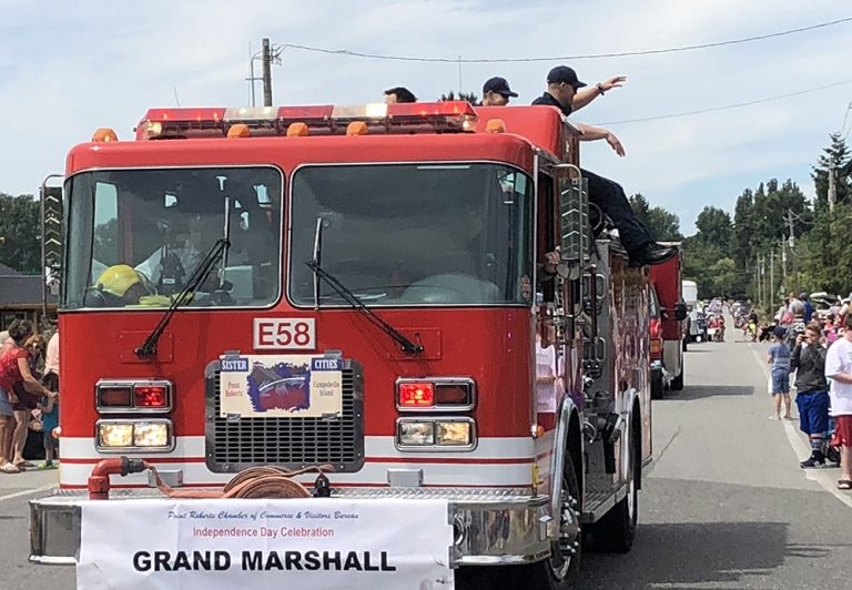 A fire truck in a parade with "Grand Marshall" banner, people on the truck waving to the crowd.