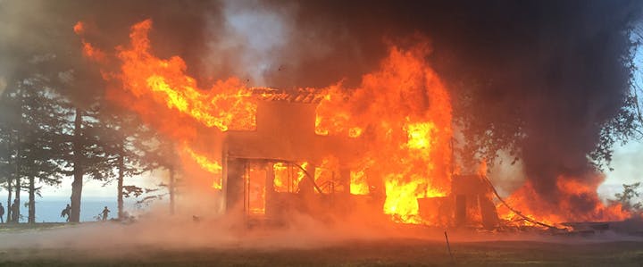 A building engulfed in flames with thick smoke rising, set against a backdrop of trees.