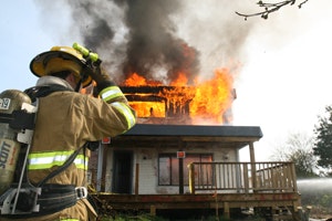 A firefighter observes a house engulfed in flames, with smoke billowing into the sky.