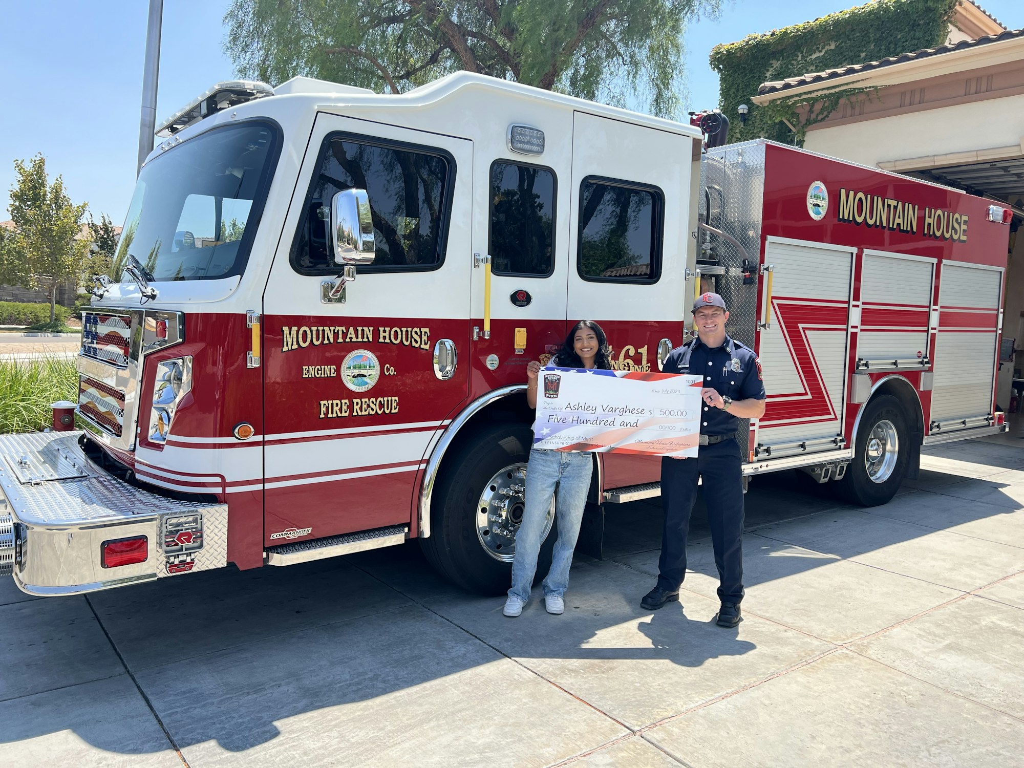 Two people standing by a fire truck, one holding a large ceremonial check.
