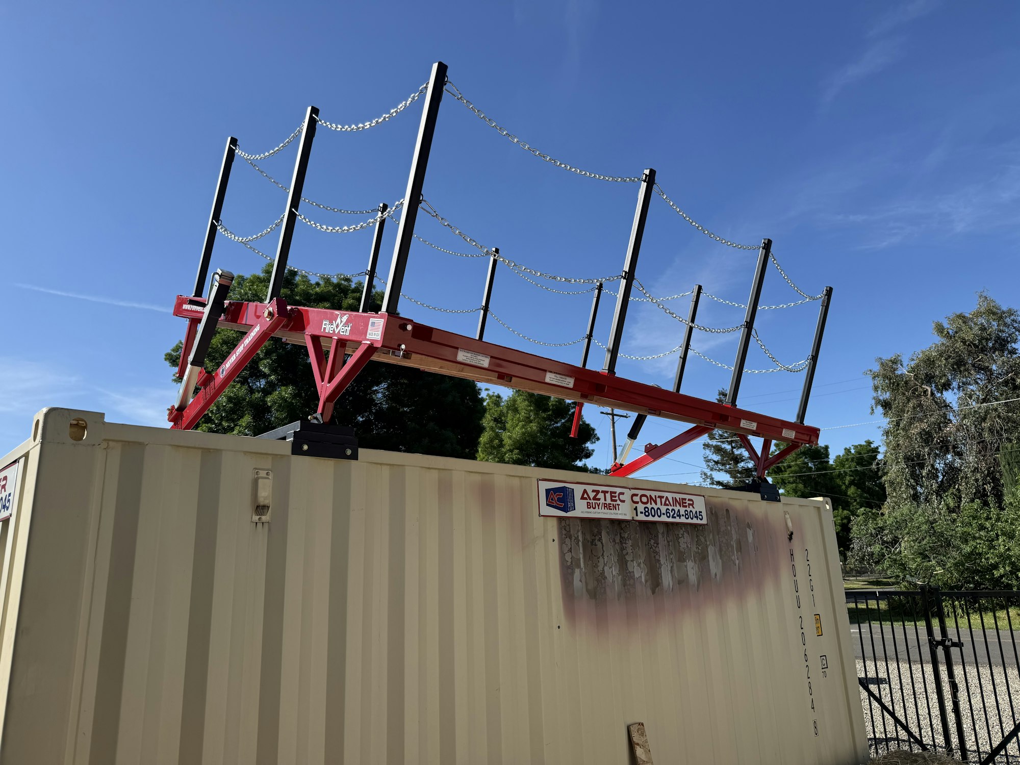 A red structure with chain links sits atop a beige shipping container under a blue sky and surrounded by trees.