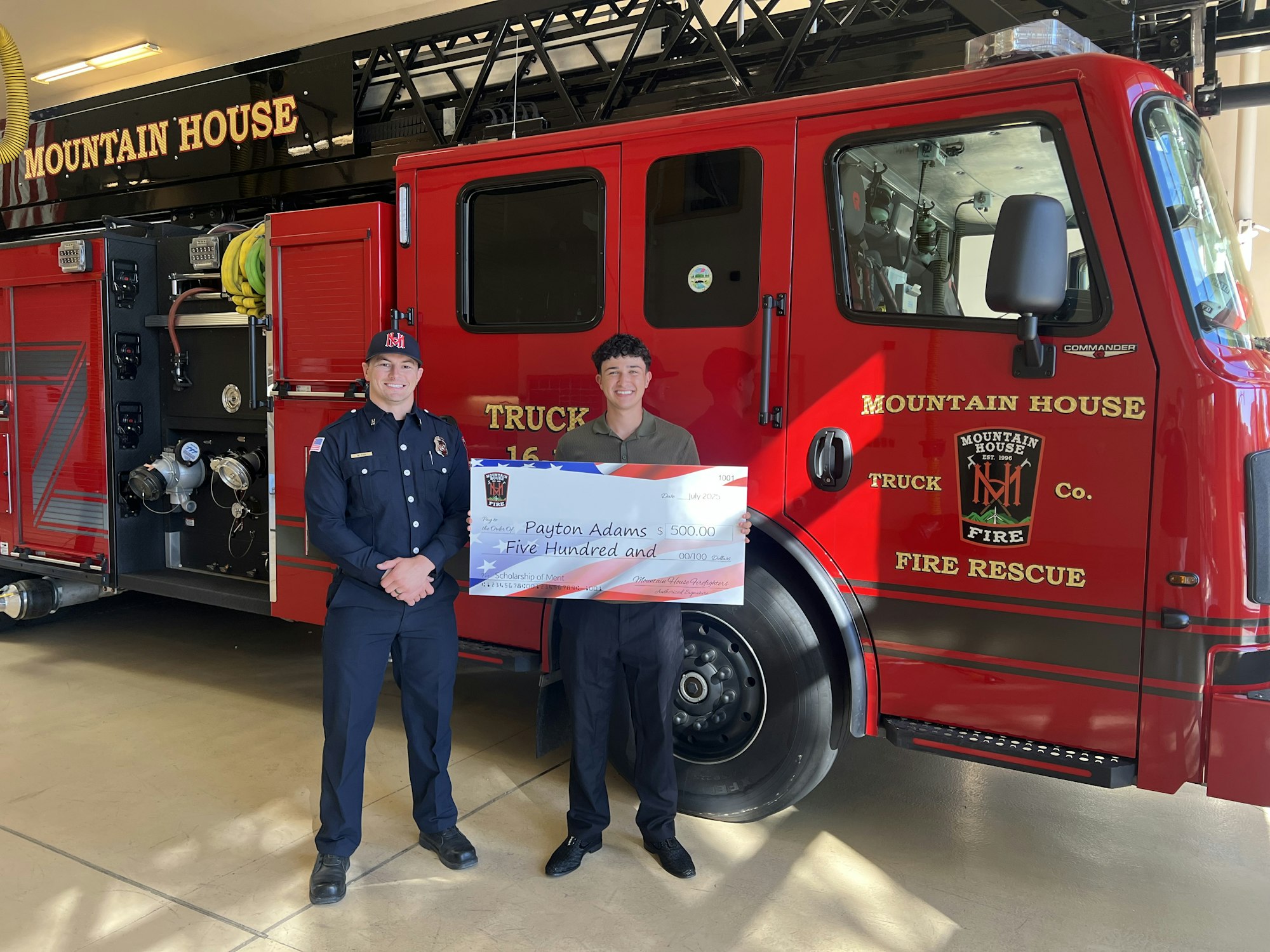 Two individuals stand in front of a "Mountain House" fire truck holding a large check for $500.