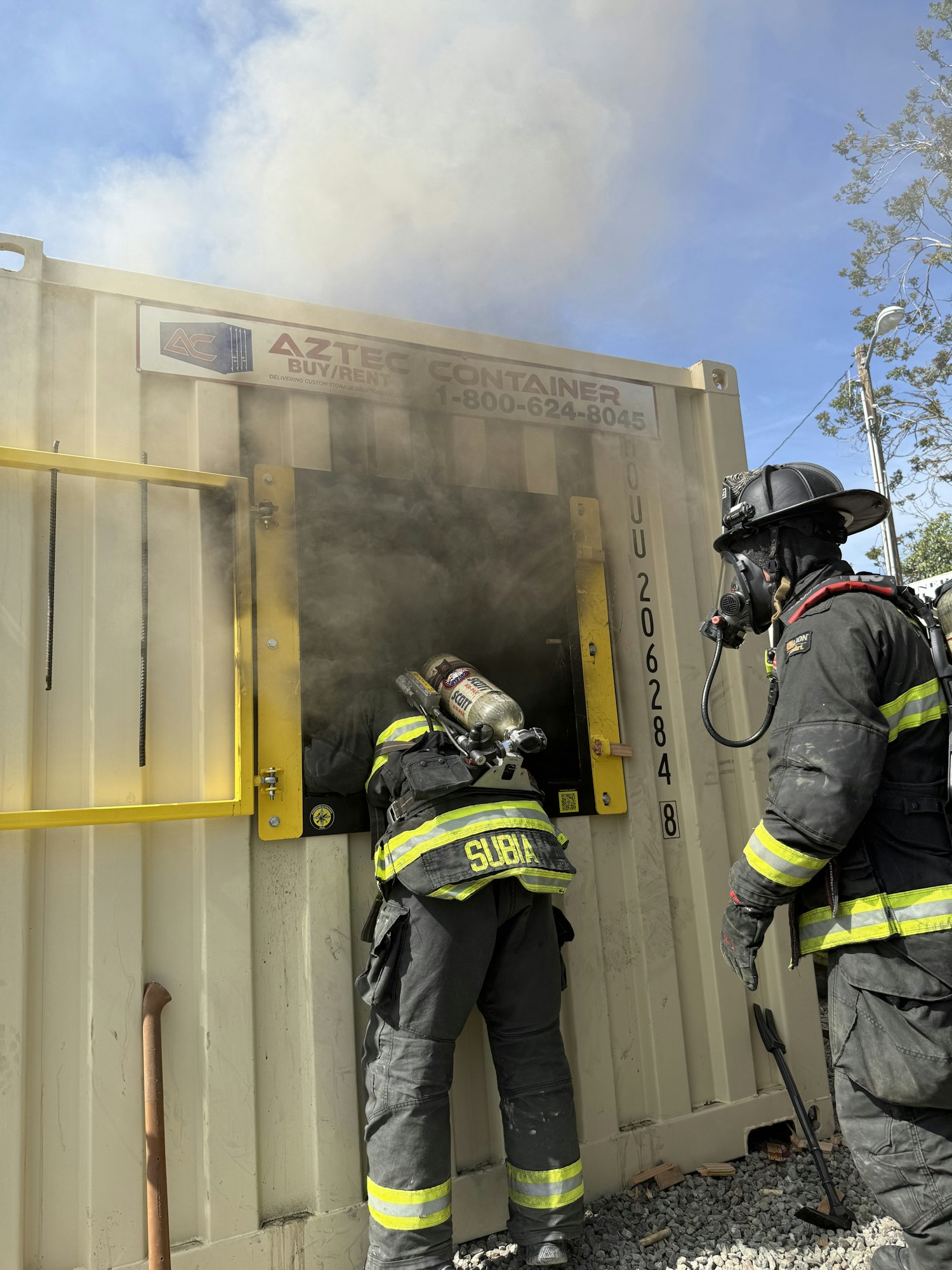 Firefighters in gear approaching a smoke-filled container, likely training for fire response or rescue operations.