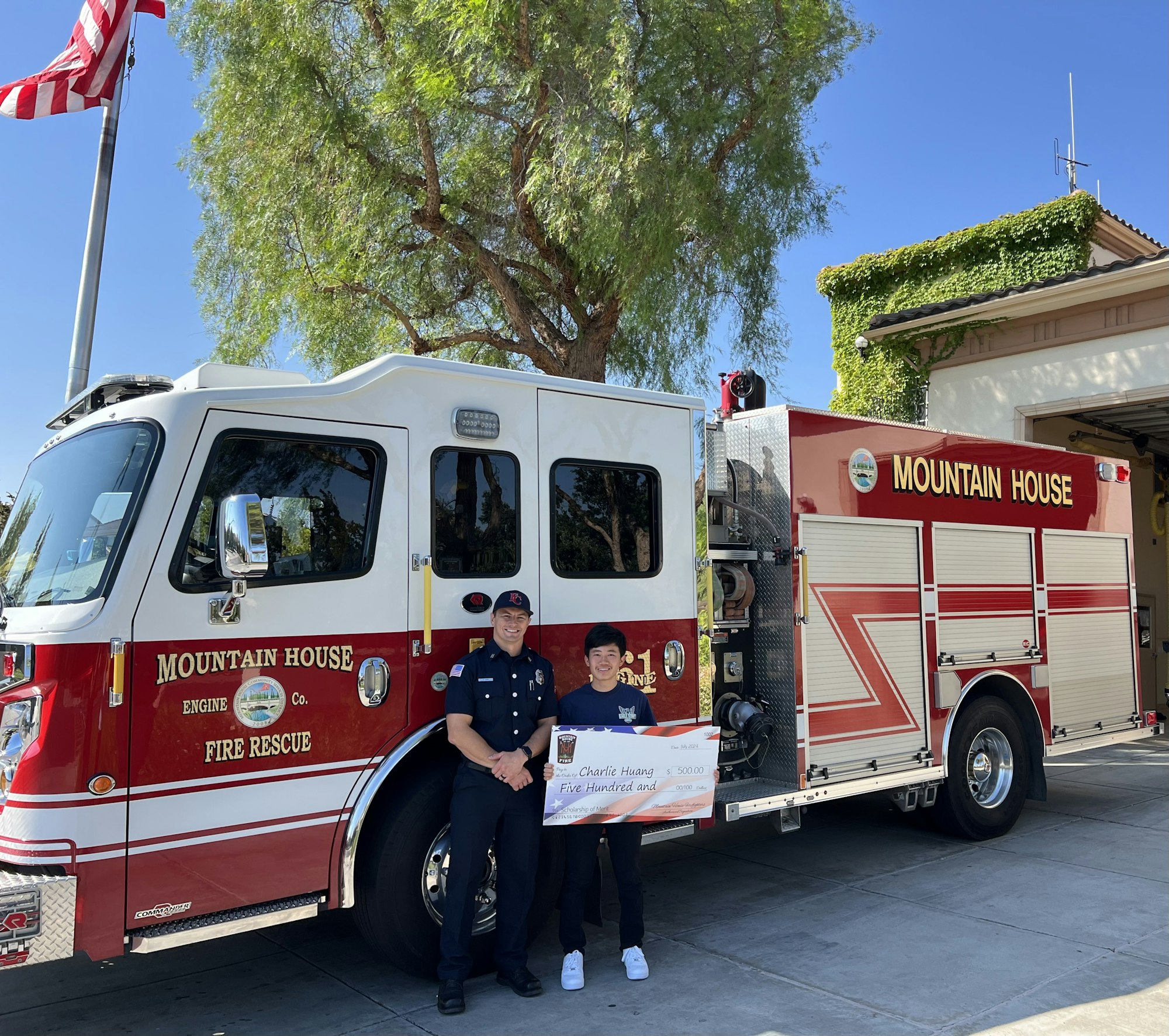 Two individuals posing with a fire truck and a donation check outside a fire station.