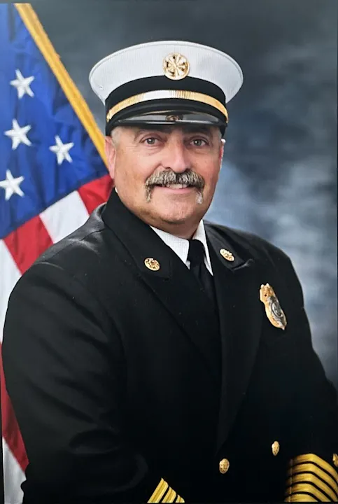 A man in a formal uniform with a peaked cap, smiling, poses in front of an American flag backdrop.