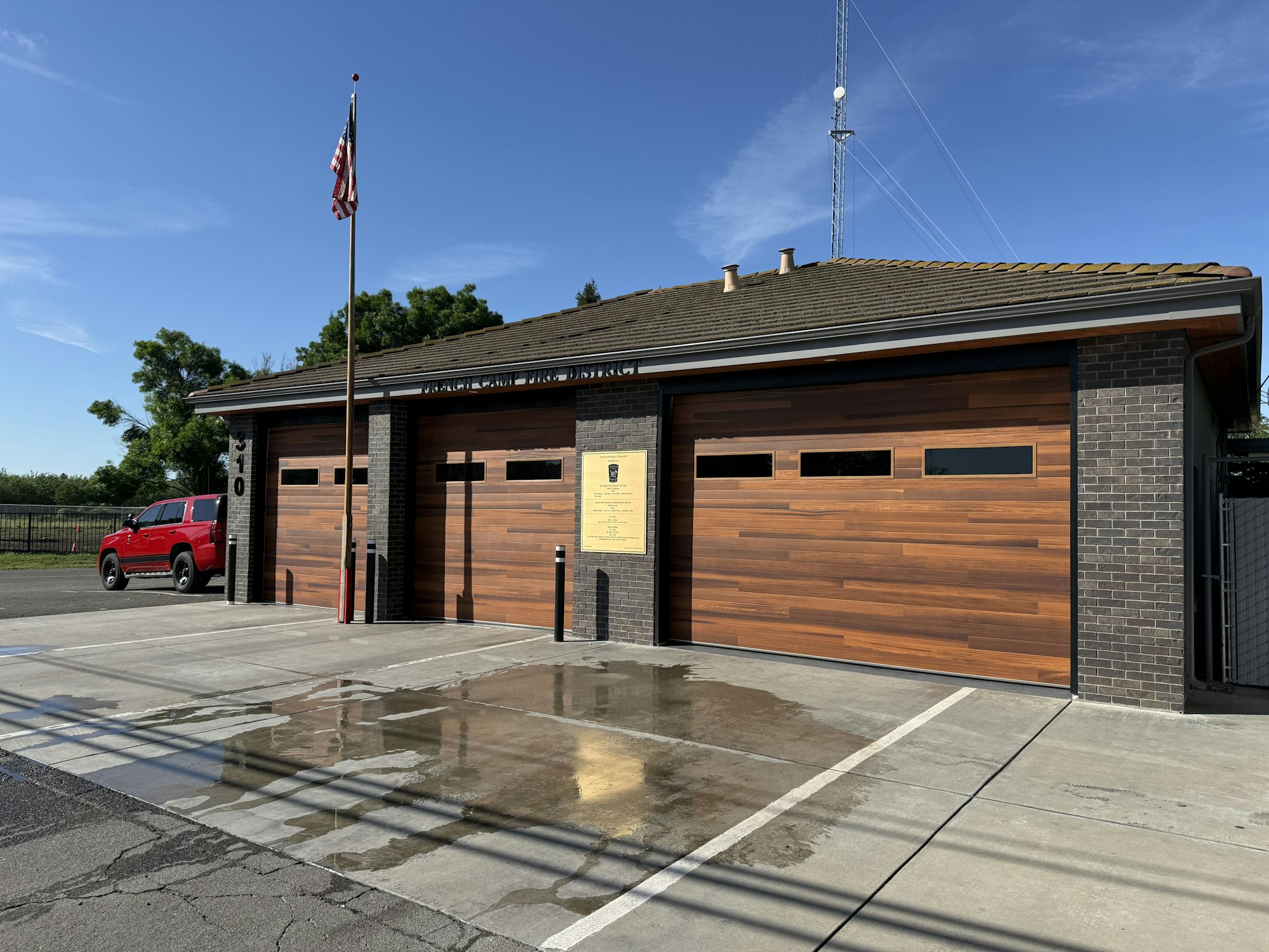 A fire station with wooden garage doors, a red vehicle parked outside, and an American flag flying in clear blue skies.
