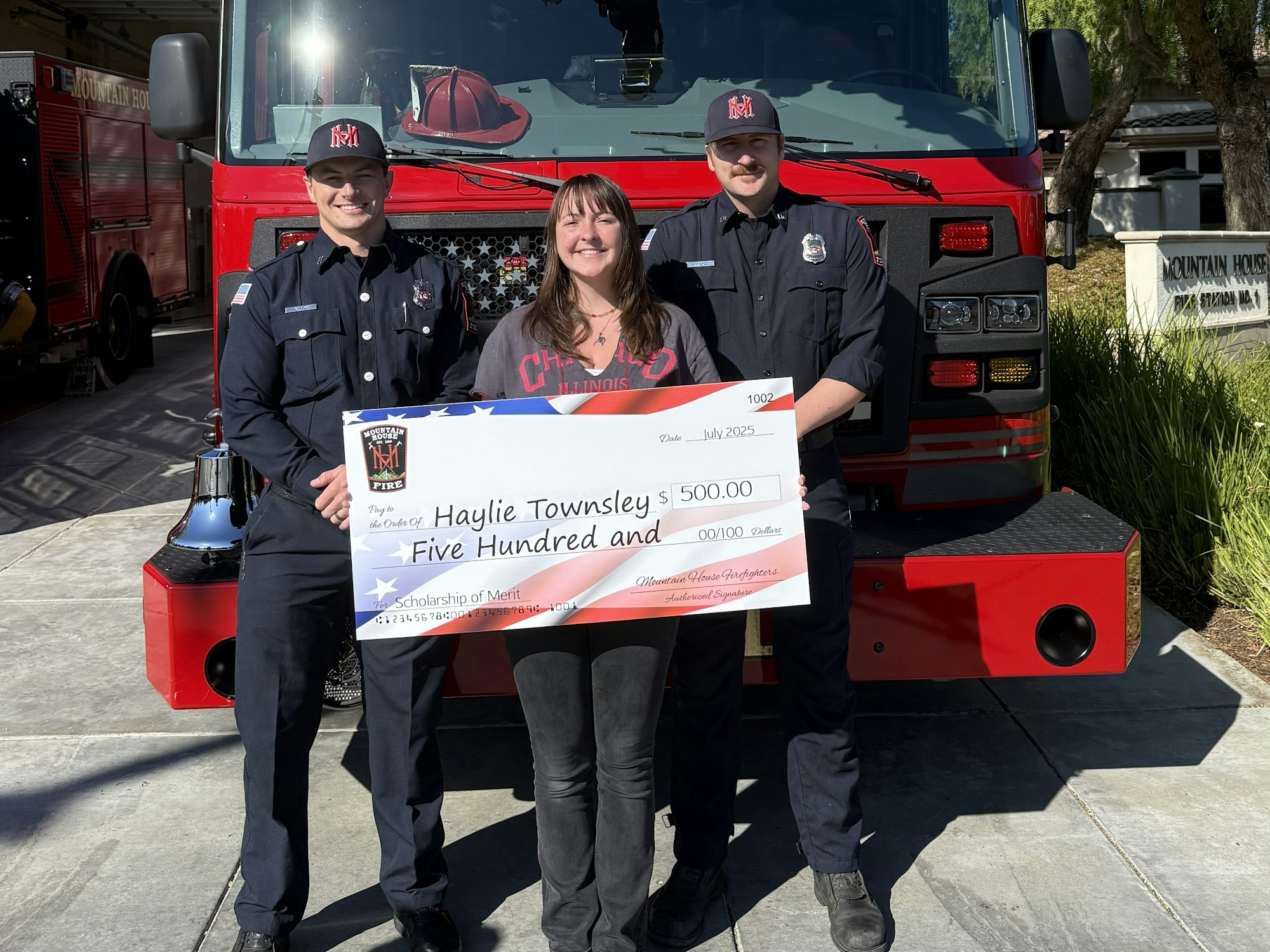 Three people holding a large check for $500 in front of a fire truck.