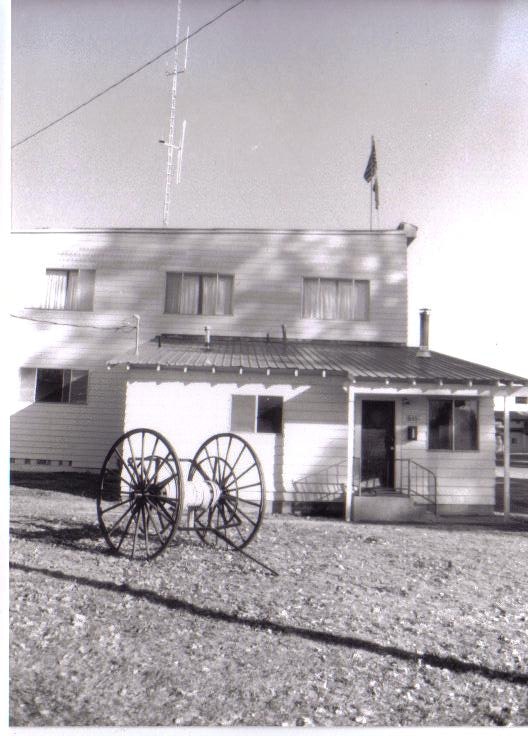 A black and white photo of a two-story building with an antenna, a flag, and old wagon wheels in the foreground.