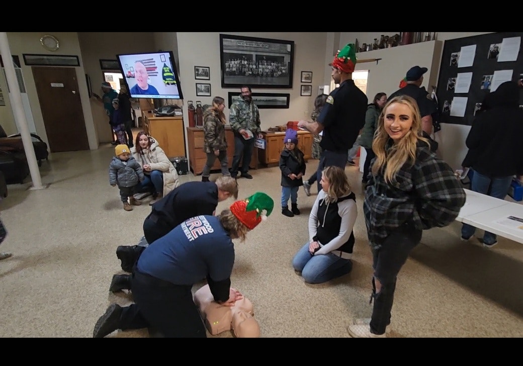 A crowded indoor gathering with people chatting and some wearing festive hats; a monitor displays a person’s face.