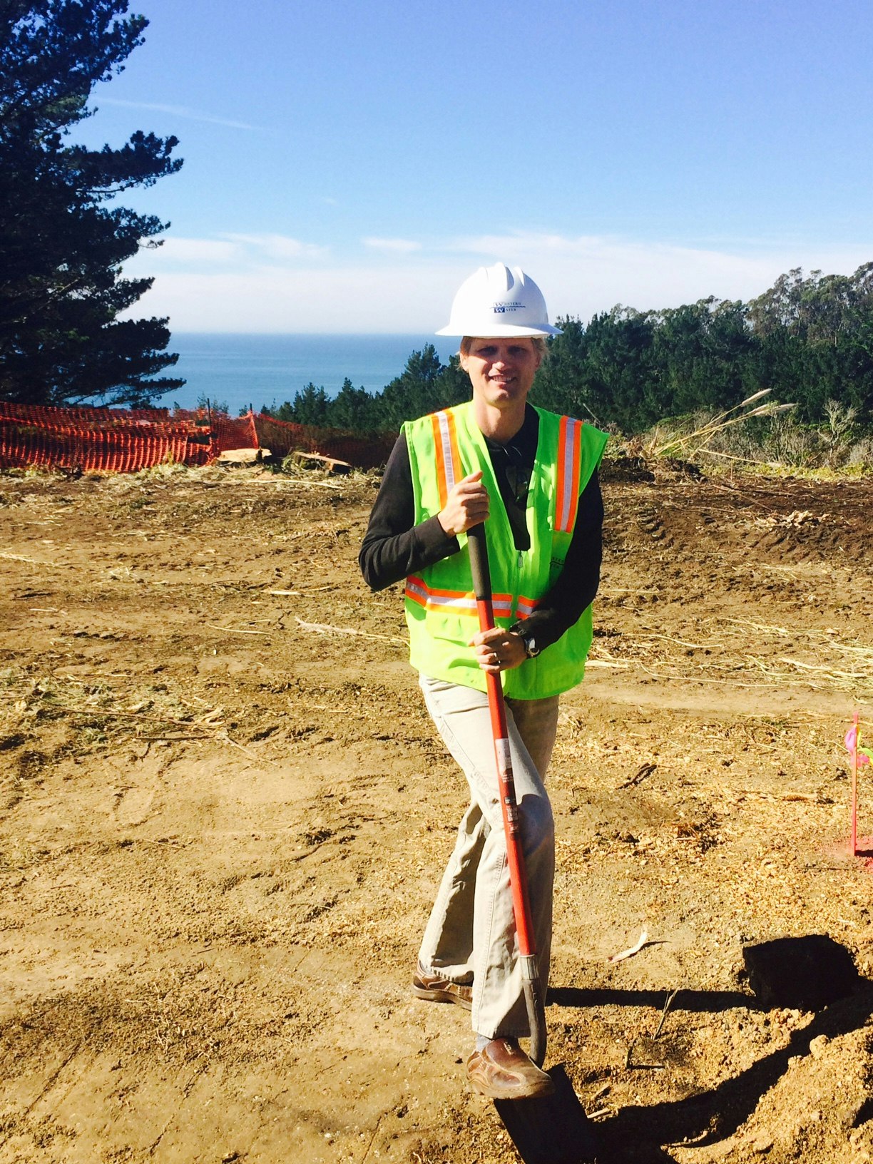 A person in a construction vest and hard hat holding a shovel on a dirt site with trees and ocean in the background.