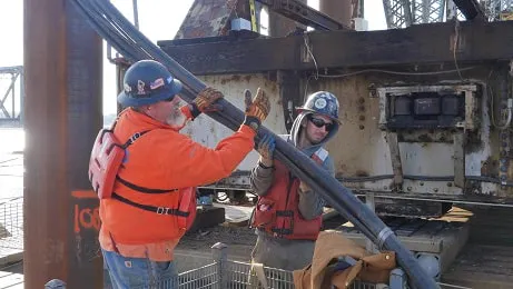 Two workers, wearing safety gear, are handling a large cable or pipe near industrial machinery on a construction site.