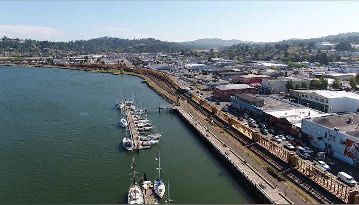 Aerial view of a coastal town with a marina, docks, parked boats, railway tracks, and surrounding buildings.
