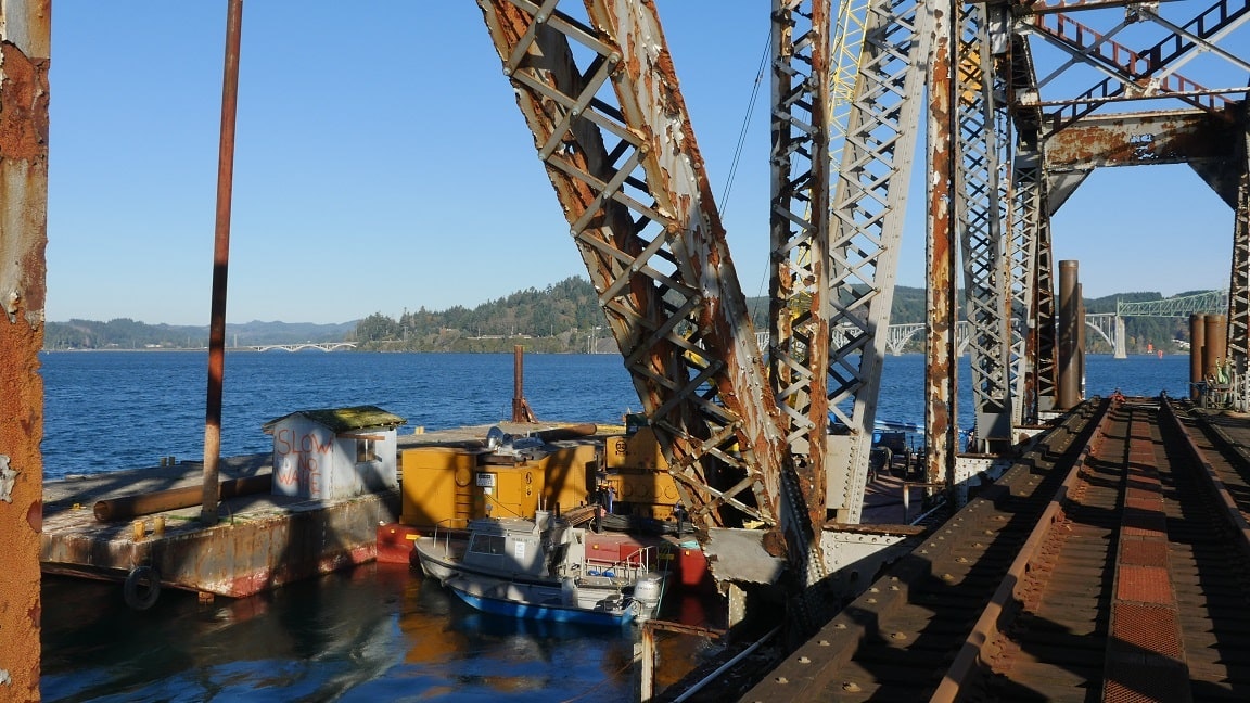 Rusty industrial structure by the water, with a small boat and yellow containers visible. Scenic view of hills and bridges in the background.