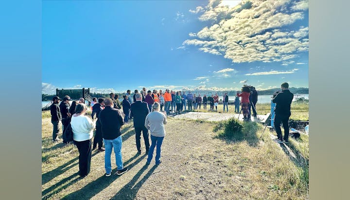 Group of people gathered outdoors, possibly for an event, with camera crew filming them, sunny sky with clouds overhead.