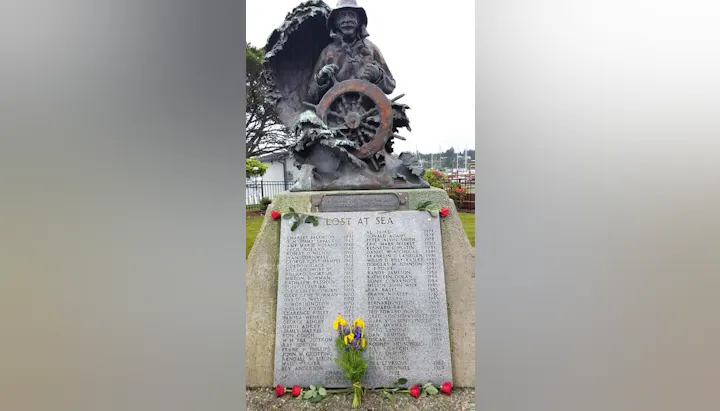 A memorial statue of a fisherman with a wheel, behind a plaque listing names of those lost at sea, adorned with flowers and roses.