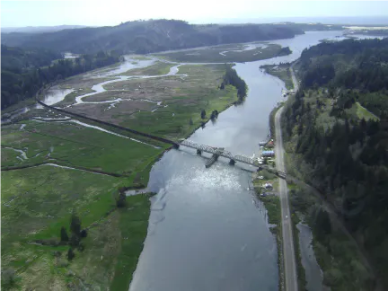 Aerial view of a river winding through green landscapes, with a bridge and road along one bank, surrounded by hills.