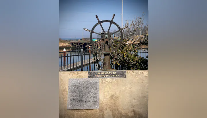 A memorial featuring a ship's wheel, honoring those from the fishing industry, with names listed on a plaque.