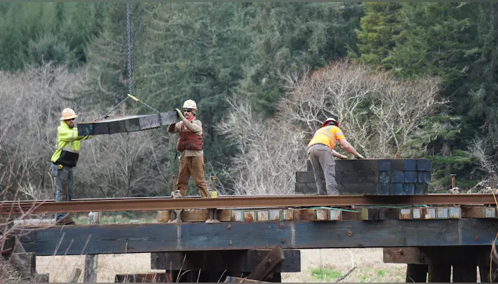 Three workers are lifting and placing heavy materials on a bridge structure surrounded by trees. Safety gear is worn.