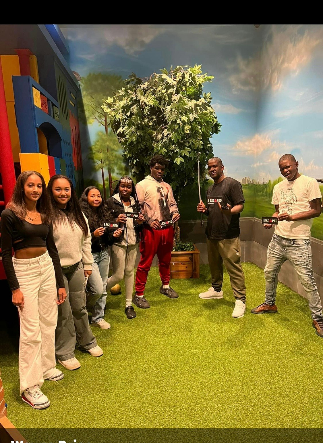 Group of people posing with props in a colorful, playful indoor setting resembling a cartoonish landscape.