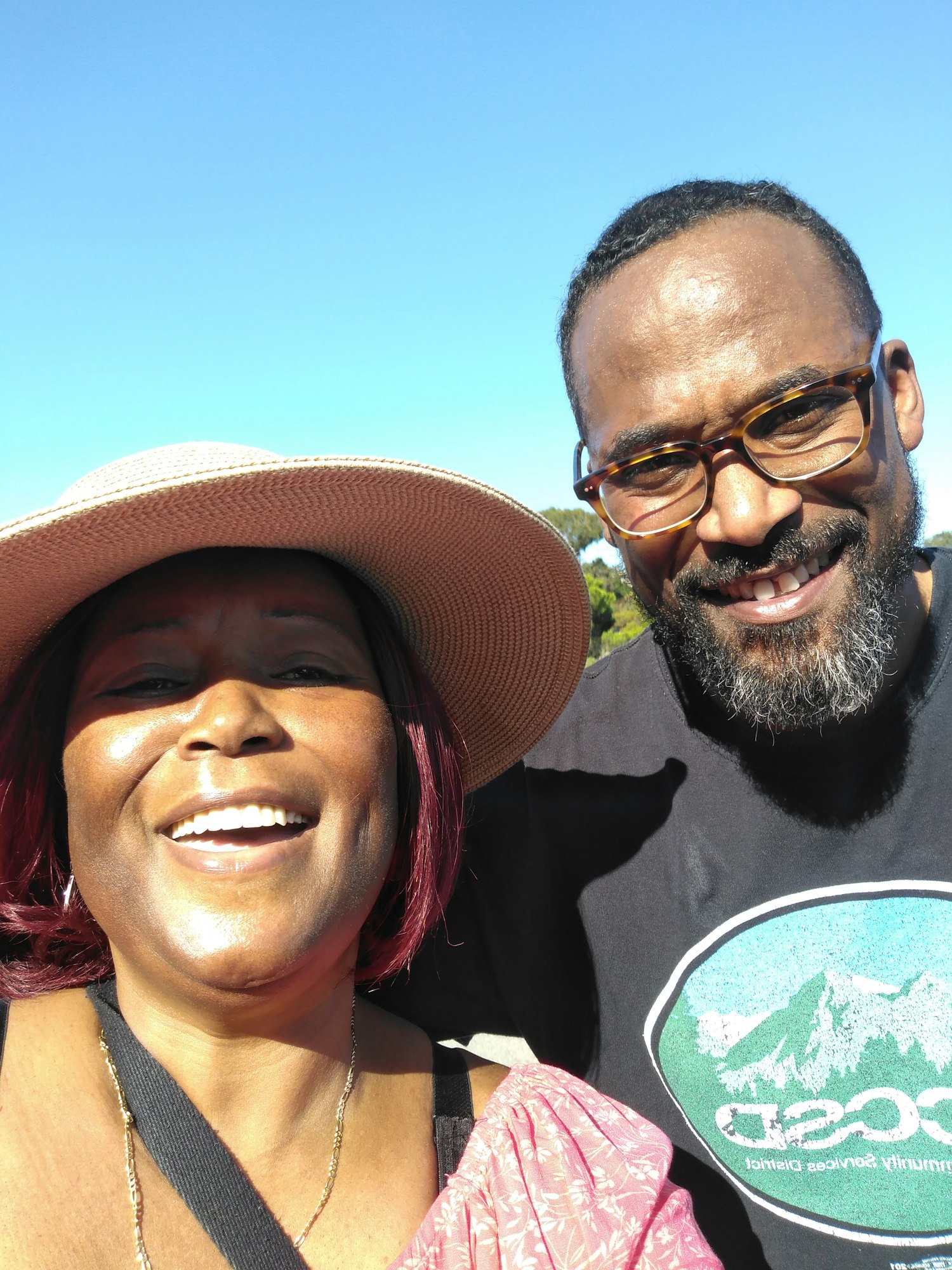 A joyful selfie of two smiling people outdoors, with a clear blue sky in the background. One wears a straw hat.