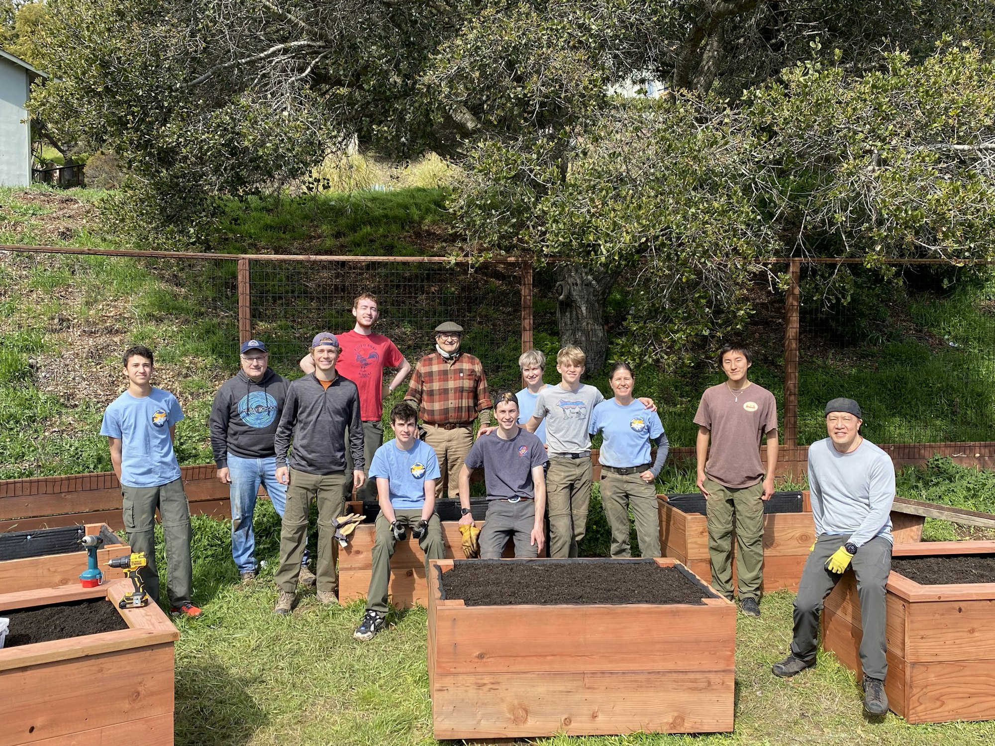 A group of people standing outdoors near wooden planters, some holding tools, with trees and grass in the background.