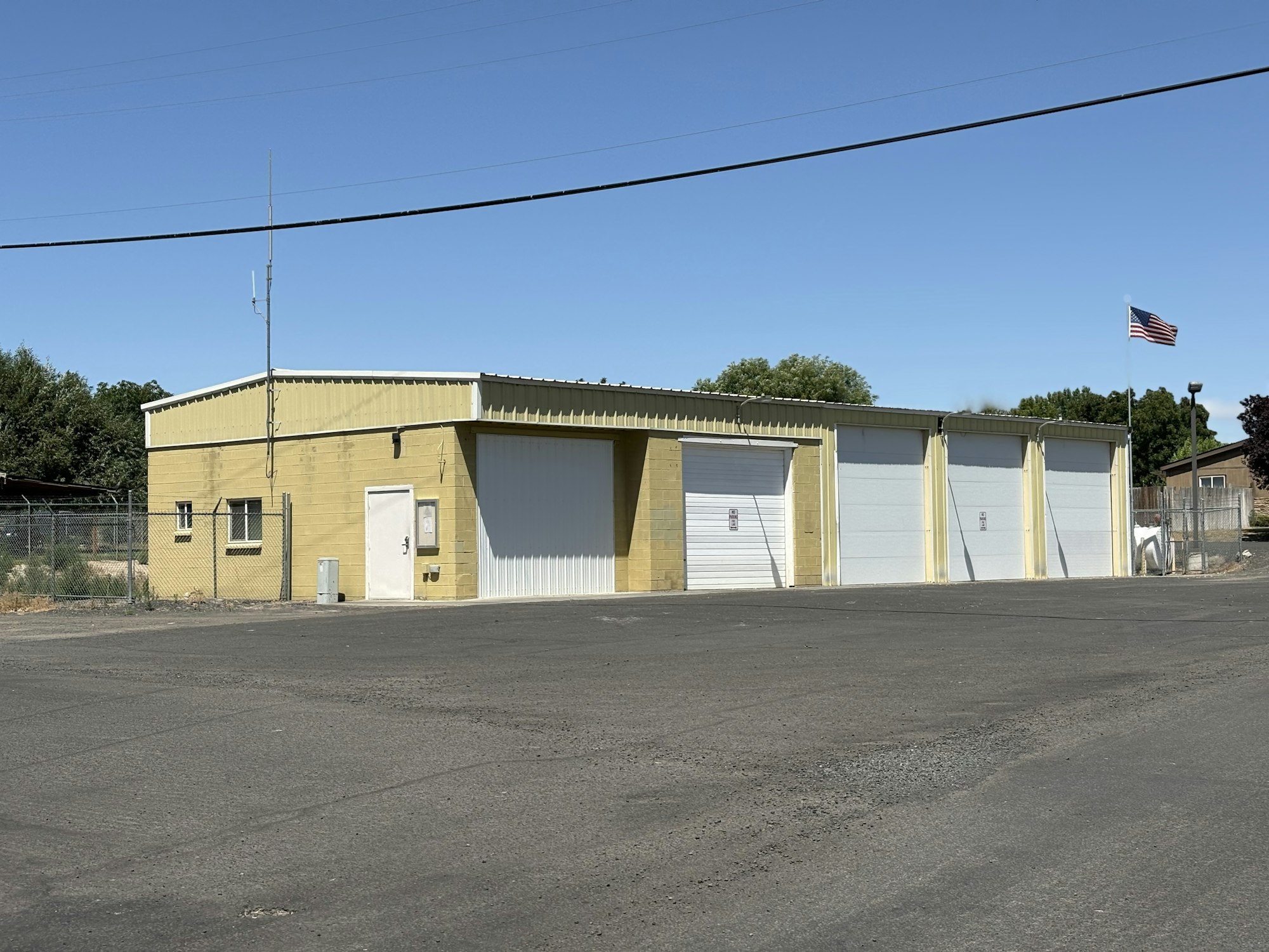 A small, yellow brick building with three garage doors, a U.S. flag, and a surrounding fence under a clear blue sky.
