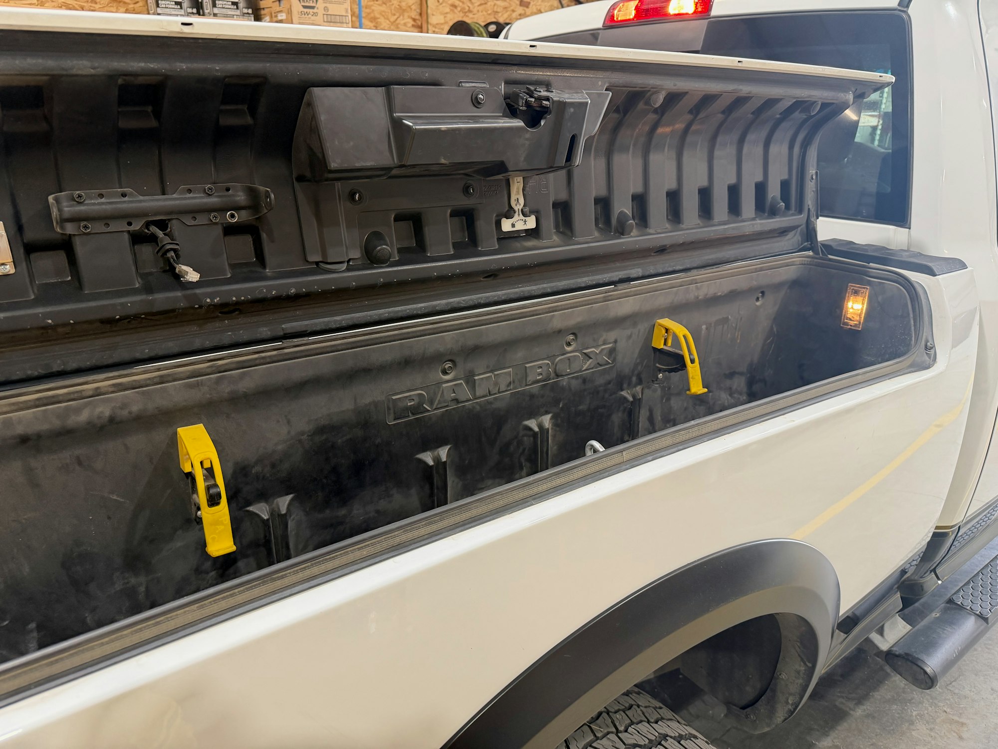 The image shows an open storage compartment in a pickup truck, featuring yellow latches, lighting, and a labeled surface.